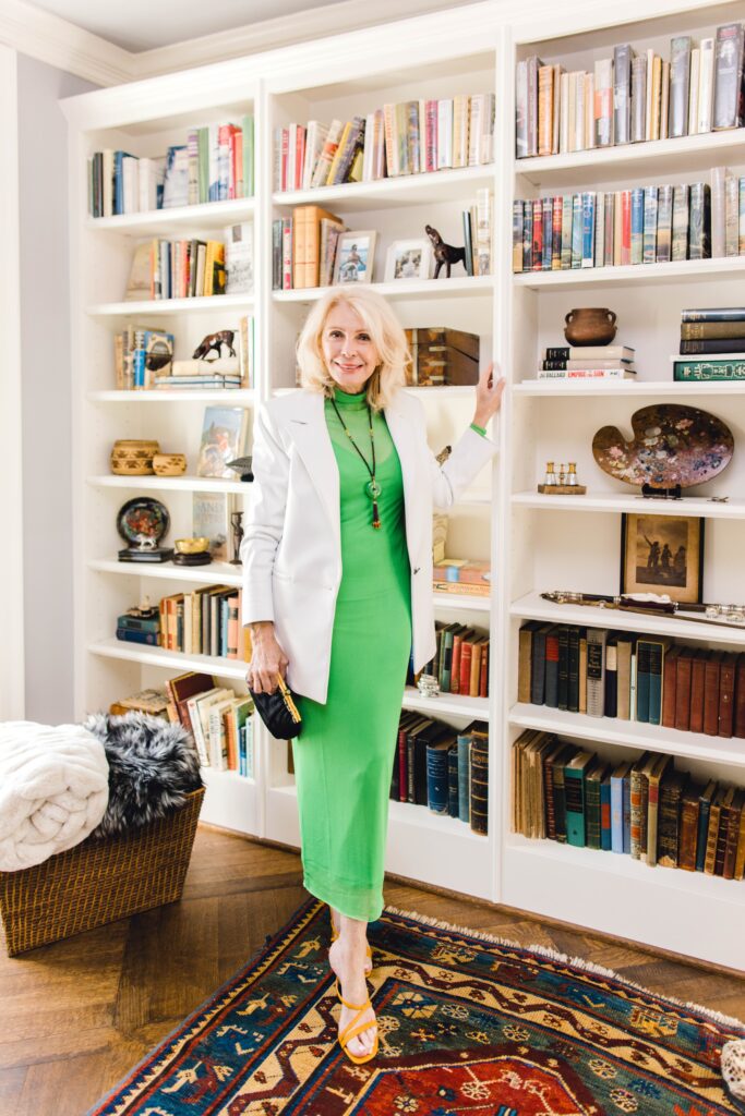 standing in front of book shelf wearing green slip dress and white leather blazer