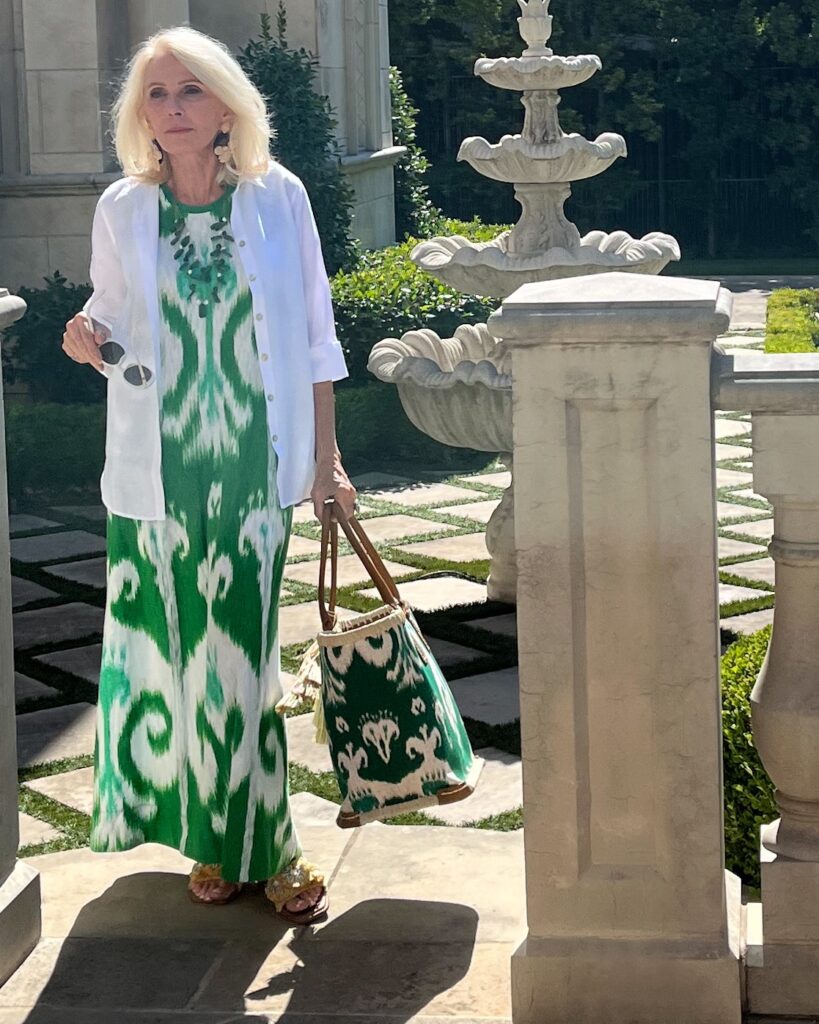Woman standing in courtyard holding white sunglasses, a tote bag and wearing a green printed dress