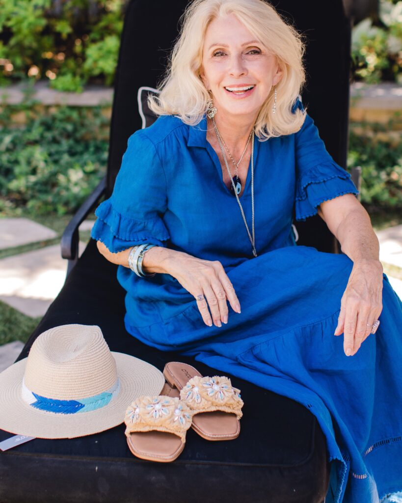 Woman in blue dress wearing jewelry sitting on chair next to straw hat and  seashell slides