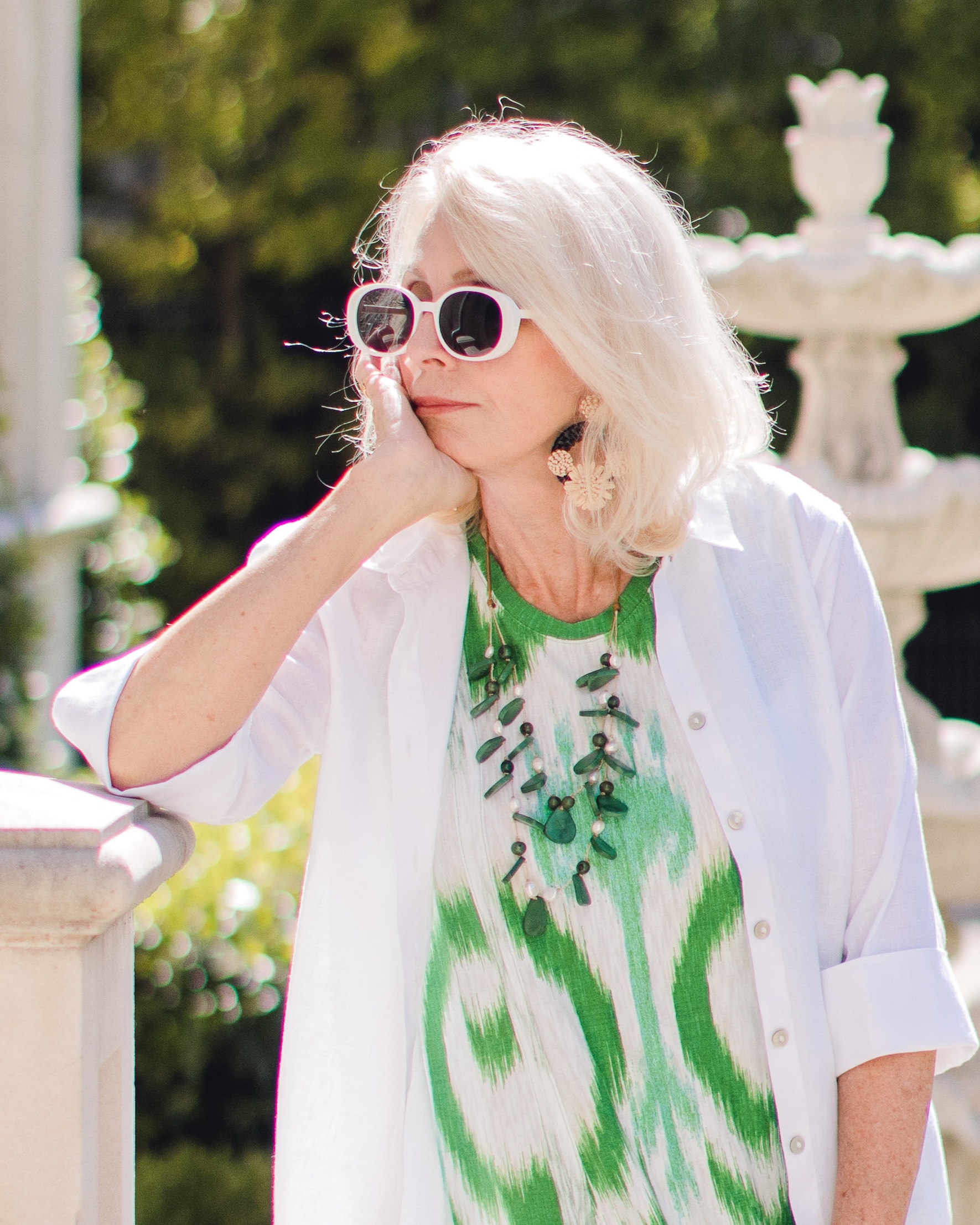 Blonde woman leaning with her hand on her chin wearing a white blouse over a green printed dress and a green necklace