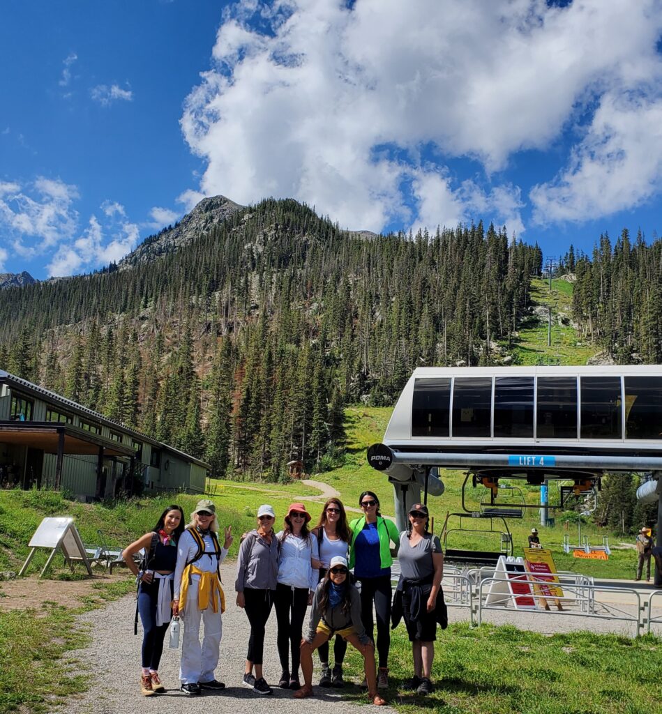 Group of women posing in front of a ski lift during summer time with a large mountain in the background