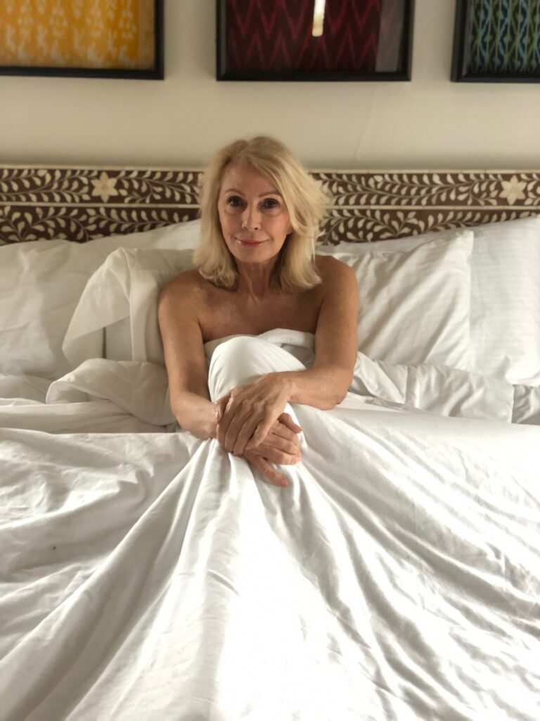 Style Beyond Age sitting in a bed with white bedding from Sleep and Glow and Lily Silk.