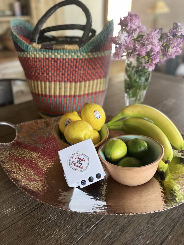 Various fruits and vegetables sitting on a metallic platter on a wooden table with flowers and a woven bag.