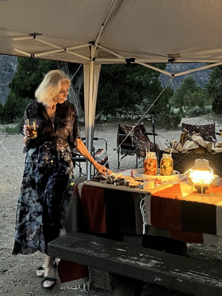 Woman in long black and white dress standing next to picnic table with lantern and canned foods underneath tent