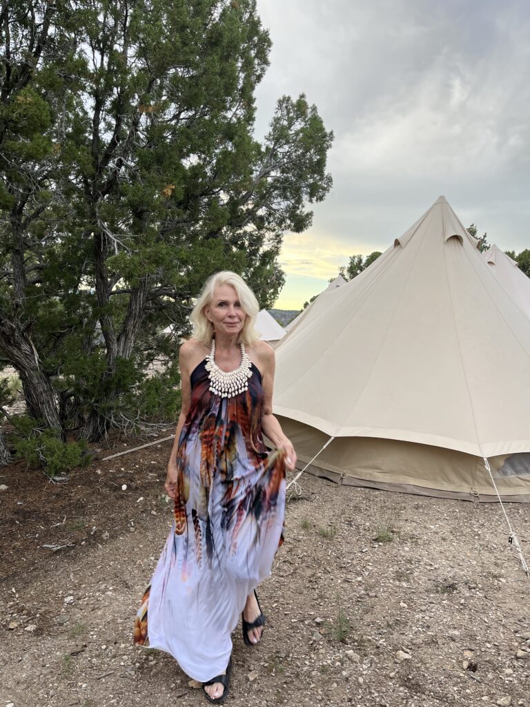 Style Beyond Age wearing white dress with colored feathers walking towards camera with tent in the background