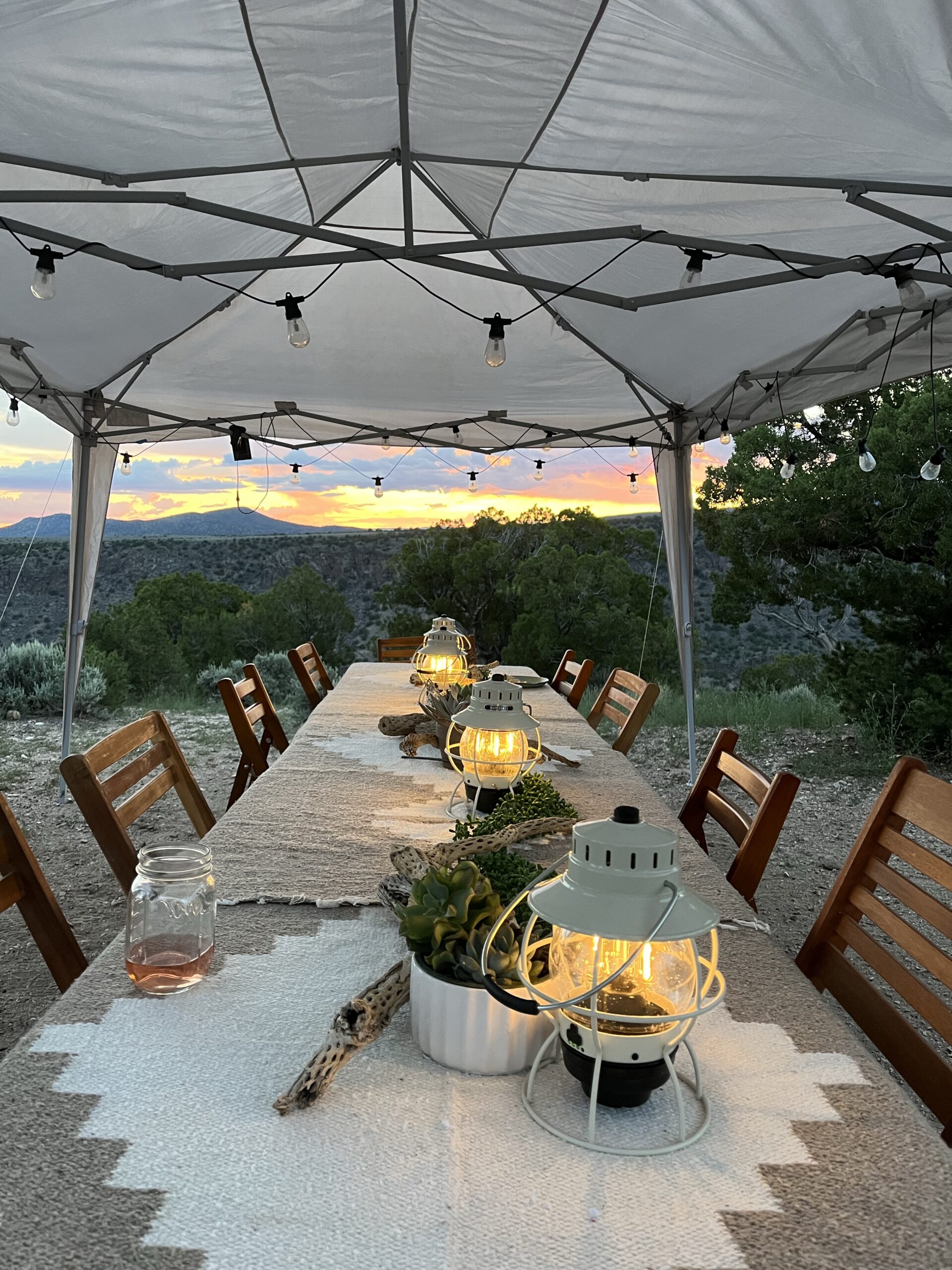 Long table under white tent with lights strung across with a scenic mountainous background and sunset