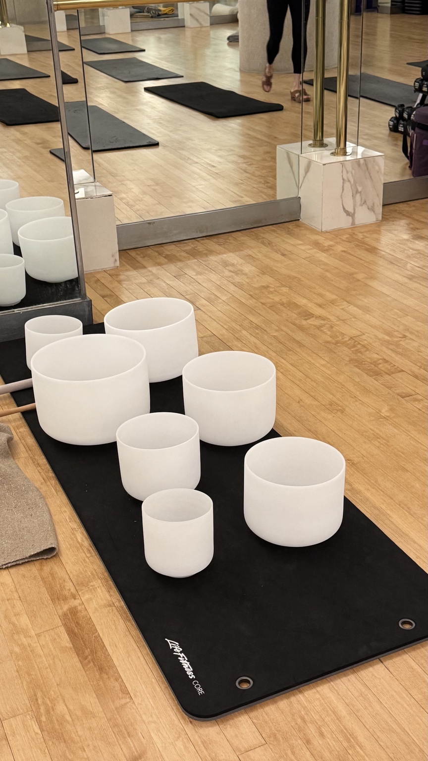 White singing bowls arranged on a black mat in the mirrored fitness studio, with mats laid out for a sound bath.